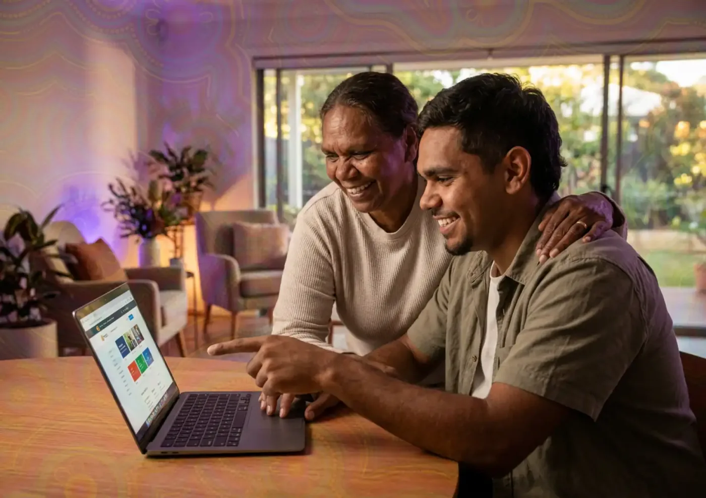 Two people seated at a table viewing a laptop together in a warmly lit room.